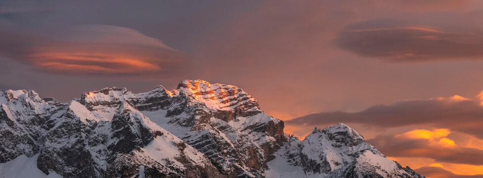 Snow-covered Brenta mountain peaks are illuminated by the warm glow of the setting sun, set against a dramatic sky filled with clouds. Adamello Brenta Natural Park,Madonna di Campiglio,Trentino,Italy