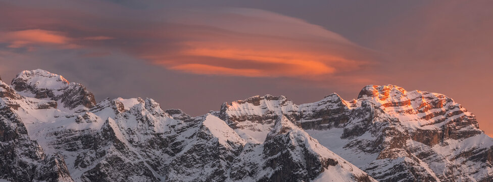 Snow-covered Brenta mountain peaks are illuminated by the warm glow of the setting sun, set against a dramatic sky filled with clouds. Adamello Brenta Natural Park,Madonna di Campiglio,Trentino,Italy