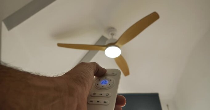 Fan's Control: A hand holds a remote control, operating a ceiling fan with wooden blades, the light on. Captured indoors, highlighting the intersection of technology and home comfort.