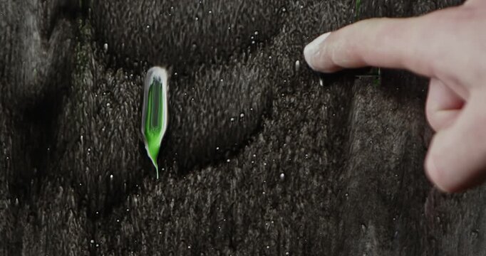 A man draws a smiley face with his finger in white and green paint on wet glass against a black background, creating an expressive abstract backdrop for creative projects.