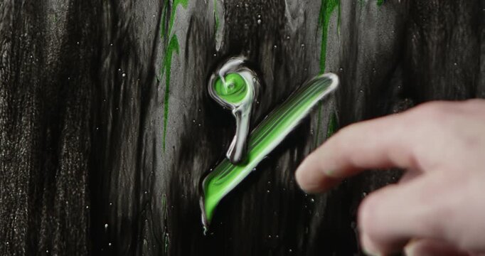 A man uses his finger dipped in white and green paint to draw a five percent symbol on wet glass against a black background, creating an expressive abstract backdrop for creative projects.