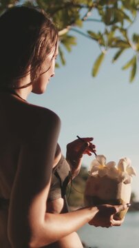Woman enjoy tropical coconut cocktail on sunset beach. Female in bikini relax under tree in soft light sea coast landscape. Female tourist on summer vacation journey. Lifestyle holiday concept