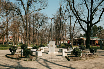 Obraz premium Urban park with benches arranged around a small monument and walking paths among tall trees. Peaceful city green space designed for relaxation, public recreation, and everyday outdoor social life.