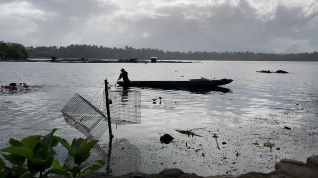 An indistinct silhouette of a fisherman is tending to fish traps while on a wooden boat in the polluted lake at sunrise.
