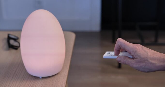 Soft Glow Ambience: A hand delicately holds a remote control, poised to adjust the settings of a stylish, egg-shaped lamp emitting a soft.