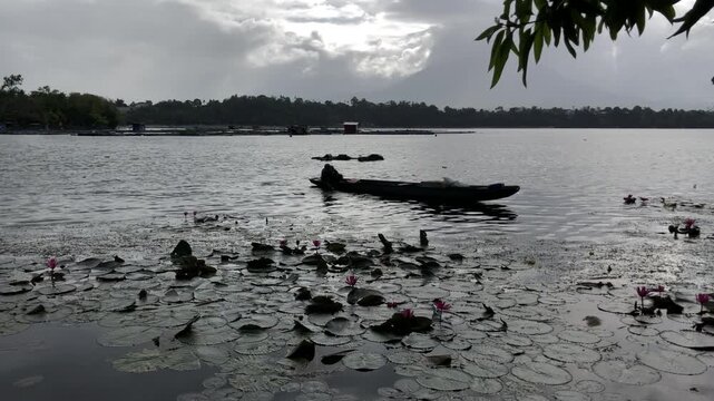 An indistinct silhouette of a fisherman is tending to fish traps while on a wooden boat in the polluted lake at sunrise.