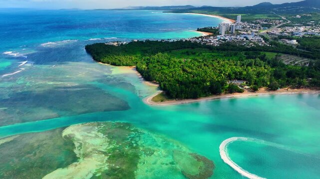 Coastal town featuring white buildings, dense green forest, sandy beaches, coral reefs, and a jet ski trail in clear blue ocean water. Aerial view of tropical coastline Puerto Rico.
