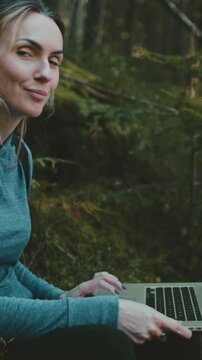 Young woman with backpack working on the laptop sitting in green forest. Freelancer working remotely. Camping, travel, outdoor adventure lifestyle.