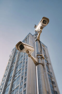 CCTV cameras and loudspeakers are mounted on a pole in front of an apartment building on a sunny summer day. The scene captures urban surveillance technology
