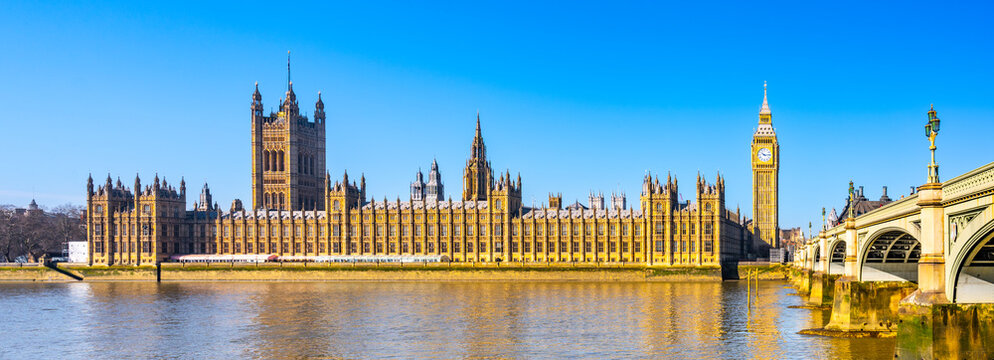 The Houses of Parliament and Big Ben along the River Thames. The sky is clear. People visit the area to admire this famous landmark.