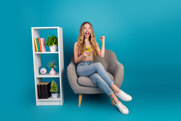 Young chic teen girl on a stylish blue set sits in a comfy chair with a phone celebrating joy