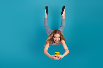 Young woman performs a handstand against a blue backdrop while using a yellow phone in a playful...