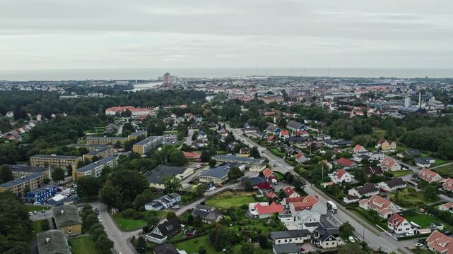 Drone shot over a quiet residential area in Falkenberg, Sweden. Tree-lined streets, detached houses and green gardens fill the foreground, with the town centre and sea visible on the horizon.