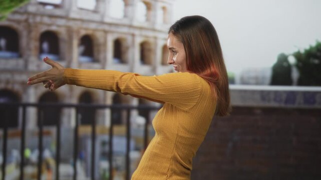 Woman redhead framing a shot with hands in finger gun pose at colosseum building, mustard sweater and side profile visible; playful travel curiosity.
