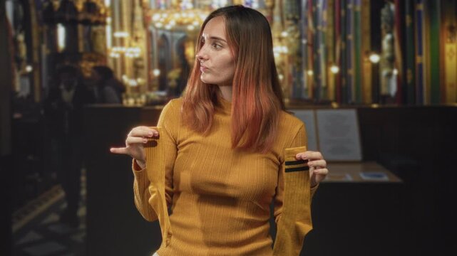 Redhead woman holds torn sleeve of mustard sweater and points finger to fabric tear, showing damage inside ornate building; quiet frustration.