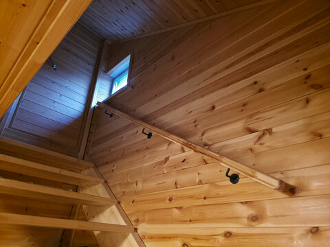 Wooden staircase in rustic house with pine wall cladding and railing. Interior design of cabin with natural light from small window. Construction and renovation concept for residential area.