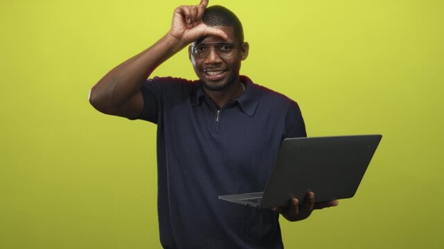 Young man wearing glasses and polo holding laptop makes l shape with hand on forehead while smiling in studio; playful confidence.