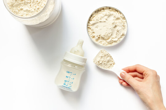 Female hand is holding powdered infant formula in a scoop and baby milk bottle with wooden teether on white background. Top view.