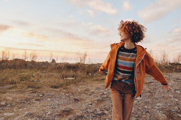 A smiling woman in a rainbow sweater and orange jacket enjoys a carefree moment outdoors at sunset...