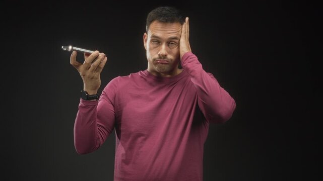 Man in magenta shirt holds smartphone at speaker with palm up and presses cheek with left hand in studio; communication frustration.