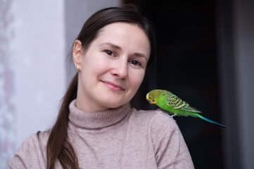 smiling brunette woman in a beige turtleneck with a bright green budgie perched on her shoulder,...