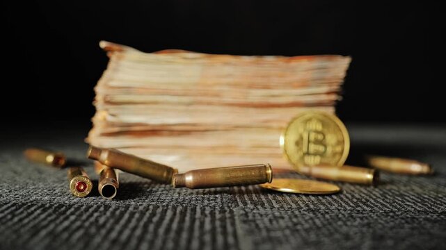 Stack of banknotes and gold bitcoin behind scattered bullets on dark floor