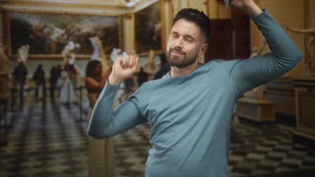 Hispanic bearded man in blue shirt holding smartphone and raising arm while dancing in ornate art museum building with marble sculptures and checkered floor; joyful.