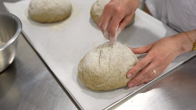 Professional baker's hands scoring a loaf of raw sourdough bread with a blade before placing it in the oven. The artisan carefully makes cuts on the floured dough resting on a baking sheet