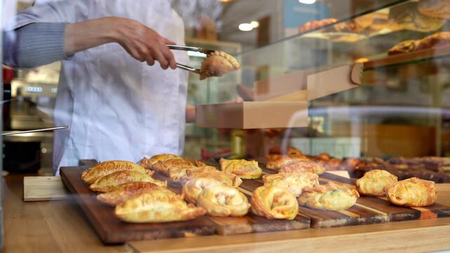 Employee in a local pastry shop carefully packing warm, savory empanadas for a customer's takeout order, placing the traditional pastries into a cardboard box with metal tongs from a wooden display