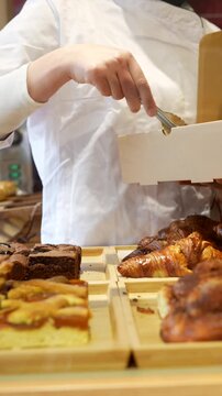 Unrecognizable pastry chef picking slices of fresh cake and sweet pastry with tongs from a display counter, putting them into a cardboard box for a customer's takeaway order