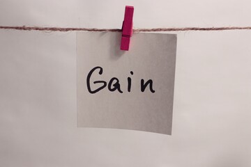 A square paper note with the word &ldquo;Gain&rdquo; written on it, attached to a string with a pink clothespin, symbolizing profit, growth, and achievement.