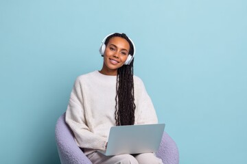 Fototapeta premium Female african adult smiling with headphones and laptop seated in a purple chair against a light blue background for remote work and study