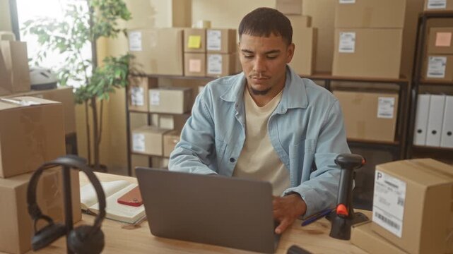 Man typing on laptop amid packing boxes and barcode scanner in studio workspace; entrepreneurial concentration.