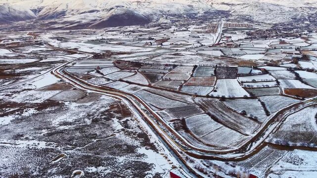 Drone aerial view of snow covered farmland stretching across a wide mountain valley. Patchwork fields, winding roads and winter terrain create a calm rural landscape beneath distant mountains.