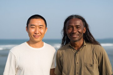 Obraz premium Asian african adult male friends smiling at beach with ocean background warm sunlight casual portrait on shore