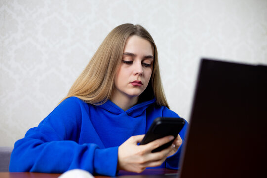 Young blonde woman uses laptop and smartphone simultaneously at home. Surrounded by digital devices, she represents modern generation immersed in technology and constant connectivity