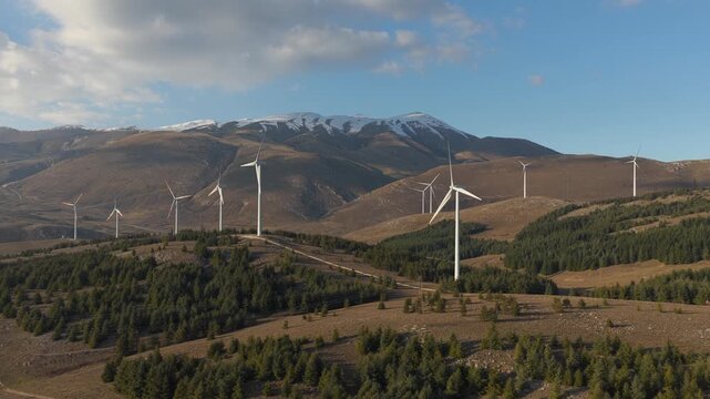 Aerial view of wind turbines dotting the rolling hills and distant snow-capped mountain peaks under a clear sky, Cocullo, Abruzzo, Italy.