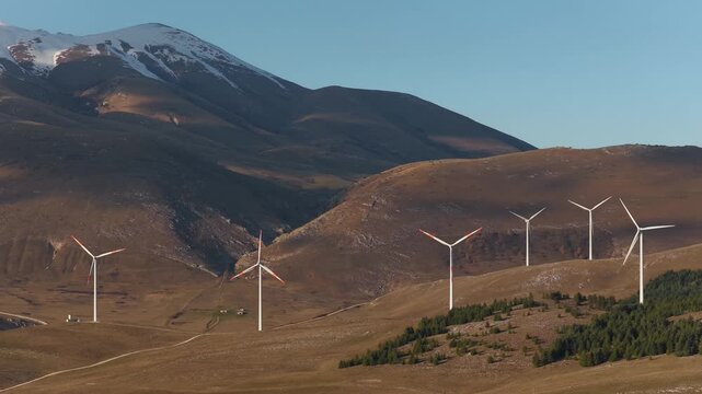 Aerial view of wind turbines standing tall against the backdrop of rolling hills and a snow-capped mountain, Cocullo, Abruzzo, Italy.