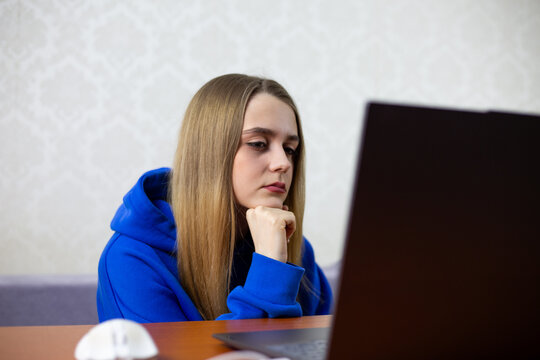 Young woman in blue hoodie sits at desk chin on fist intently watching Football World Cup. National team match live stream on laptop screen focused expression indoor home setting