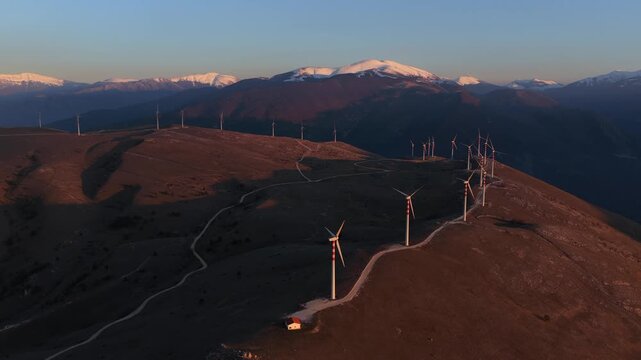 Aerial view of wind turbines aligned along a hilltop, contrasting with the snow-capped mountains in the background, Cocullo, Abruzzo, Italy.