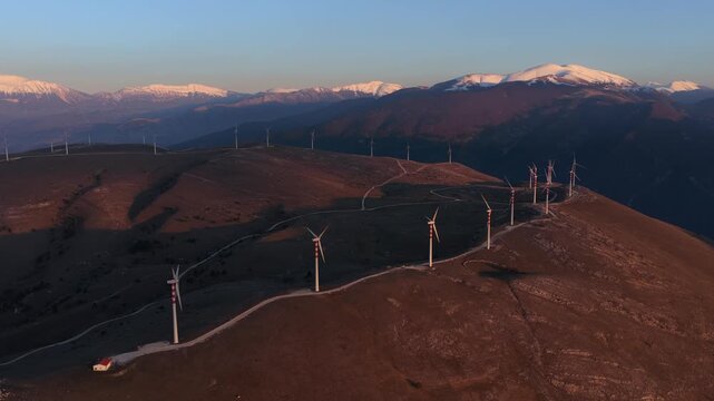 Aerial view of wind turbines standing tall on a mountainous landscape in warm light, creating a striking contrast with the distant snow-capped peaks, Cocullo, Abruzzo, Italy.