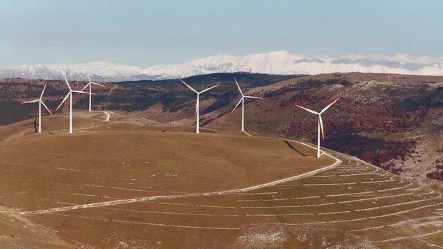 Aerial view of wind turbines stand tall against the backdrop of snow-capped mountains, creating a striking contrast, Cocullo, Abruzzo, Italy.