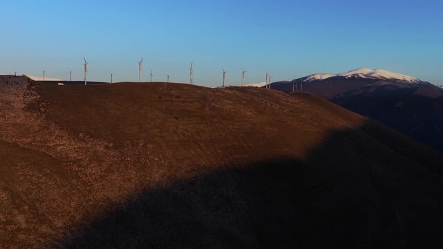 Aerial view of wind turbines standing tall and still on brown hills with snow-capped mountains in the background, Cocullo, Abruzzo, Italy.