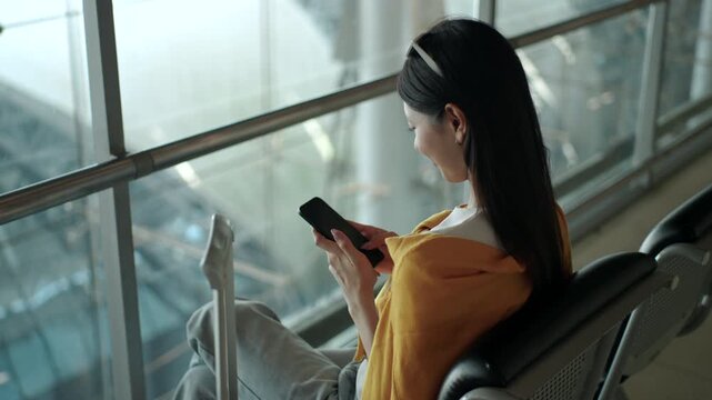 Happy young solo  woman traveler using smartphone and holding boarding pass while sitting on airport waiting chairs at the terminal, enjoying travel time before departure. Modern travel and travel ins