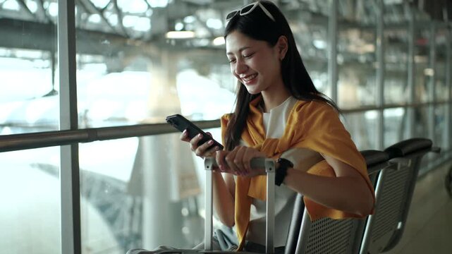 Happy young solo  woman traveler using smartphone and holding boarding pass while sitting on airport waiting chairs at the terminal, enjoying travel time before departure. Modern travel and travel ins