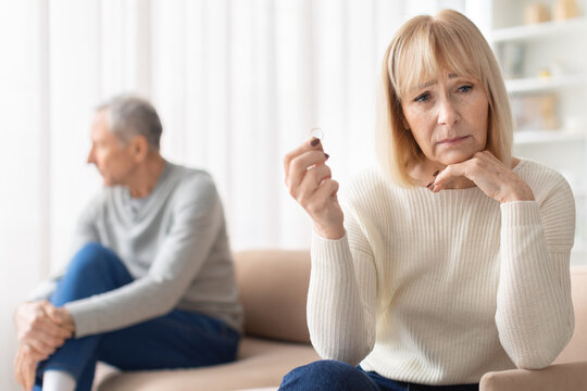 Upset senior woman holding wedding ring while sitting apart from husband on sofa. Mature couple experiencing relationship conflict representing divorce, separation and emotional distress
