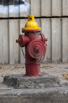 A vintage-style fire hydrant with a striking red-and-yellow colour scheme stands on a concrete base near a corrugated metal wall. The hydrant features a yellow top marked with &bdquo;DBO&ldquo; in white, secured 