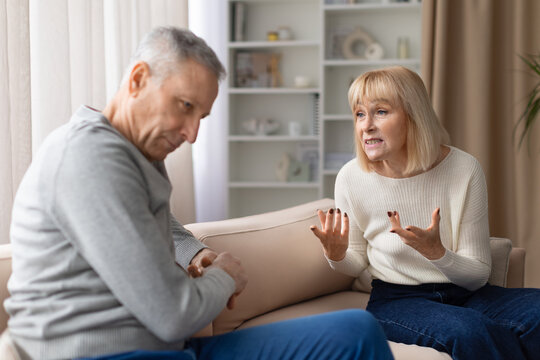 Senior couple experiencing relationship conflict while talking on sofa at home. Mature woman gesturing emotionally while man looks down during serious discussion about personal problems