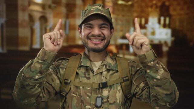 Man in camouflage uniform with dog tag smiling while pointing finger up in wooden pews of church; faith devotion service hope.
