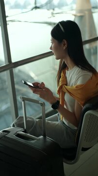 Unhappy young solo woman traveler sitting on airport waiting chairs, holding smartphone and boarding pass while waiting for delayed flight, feeling bored and frustrated at airport terminal.
